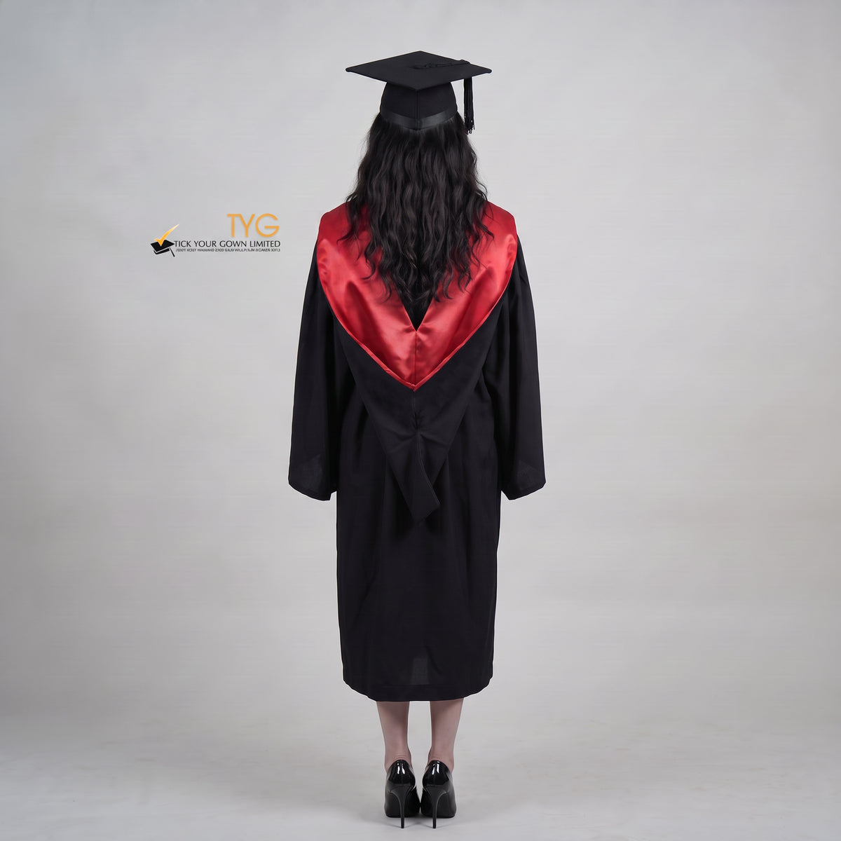 A back view woman wearing a University of Edinburgh bachelor graduation gown and a black mortarboard cap stands against a light gray studio background.
The gown has a red and black draped hood, and she is wearing a white collared shirt underneath.
