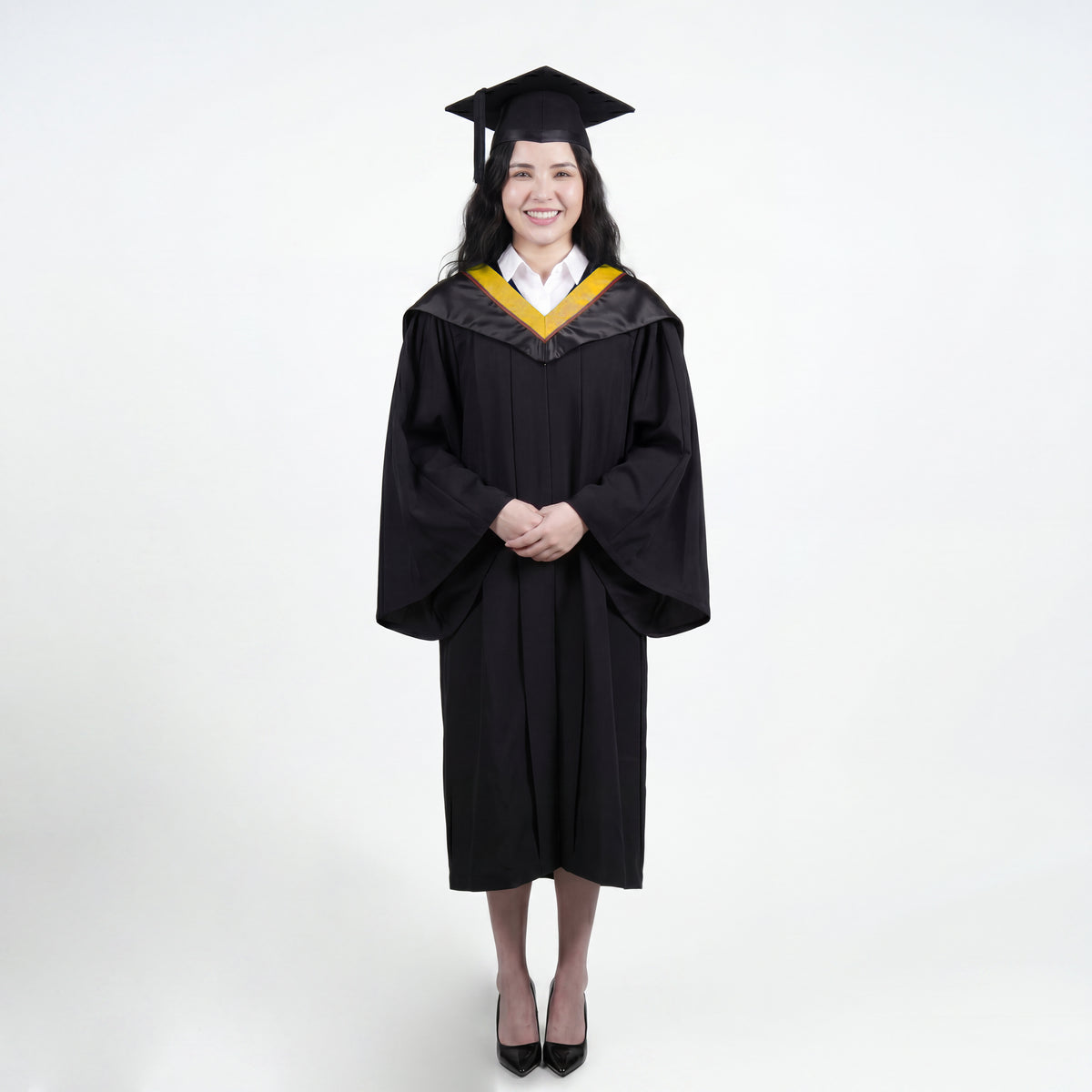 A woman wearing a black graduation gown and matching mortarboard cap stands against a light gray studio background.
The gown has a wide collar with gold and brown trim accent hood, and the model is posed with hands clasped in front.