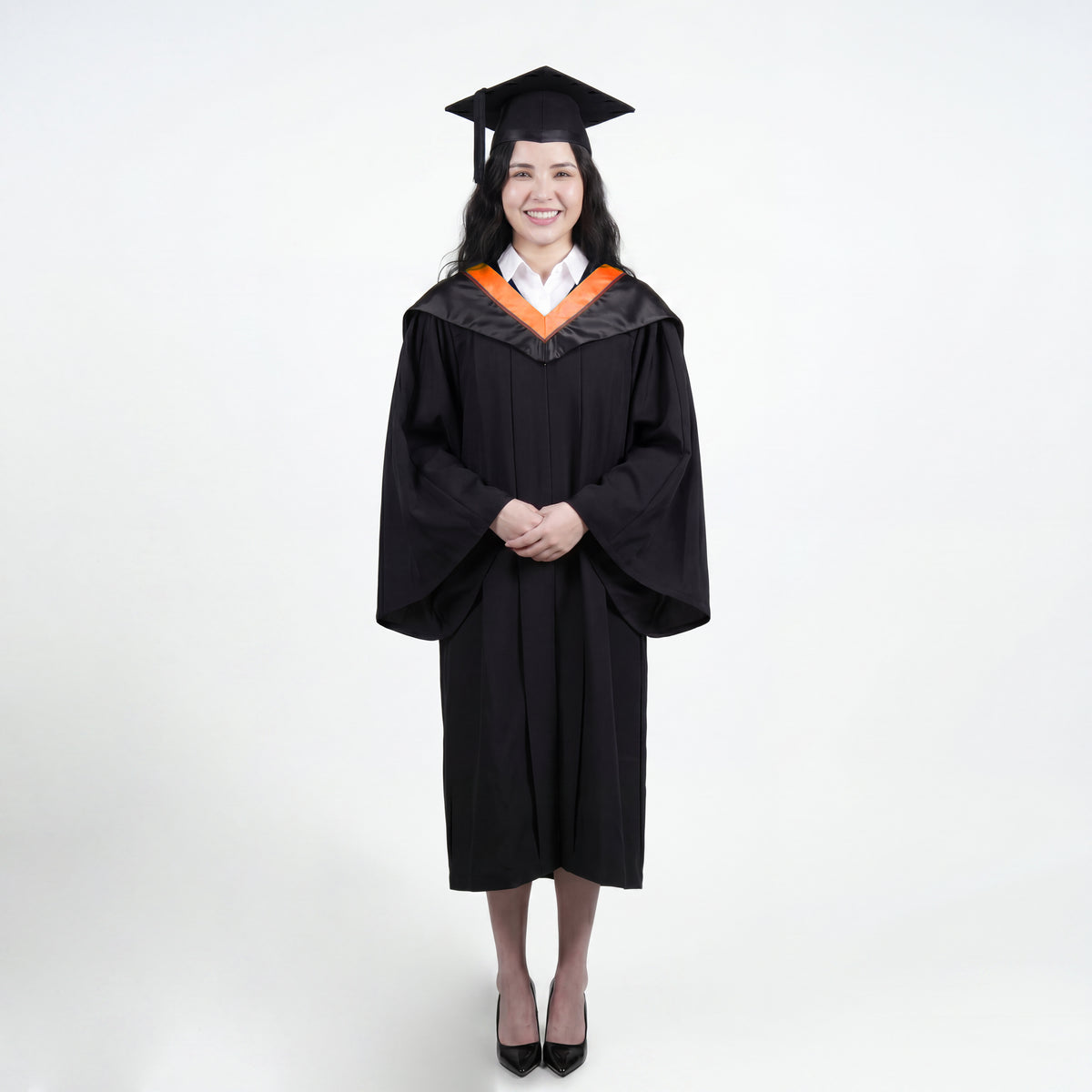 A woman wearing a black graduation gown and matching mortarboard cap stands against a light gray studio background.
The gown has a wide collar with orange and brown trim accent hood, and the model is posed with hands clasped in front.