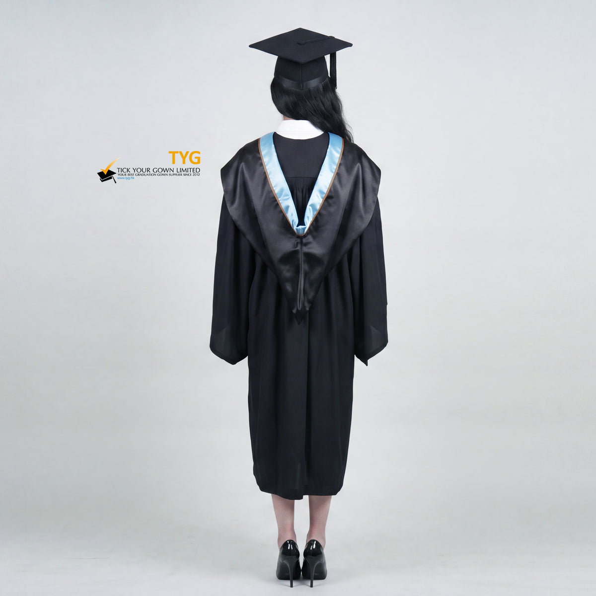 A back view of a woman wearing a black graduation gown and matching mortarboard cap stands against a light gray studio background.
The gown has a wide collar with copper and brown trim accent hood, and the model is posed with hands clasped in front.