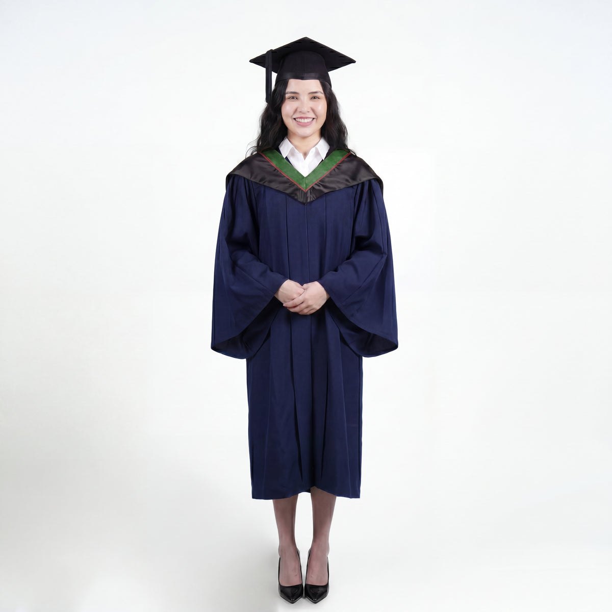 A woman wearing a navy blue graduation gown and matching mortarboard cap stands against a light gray studio background.
The gown has a wide collar with green and brown trim accent hood, and the model is posed with hands clasped in front.