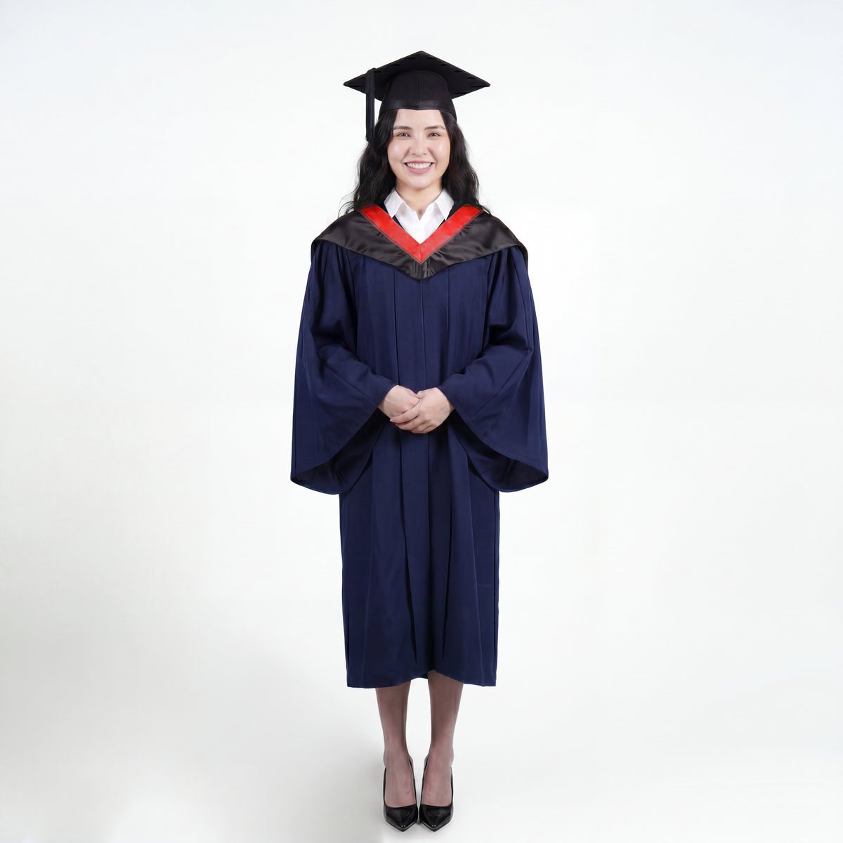A woman wearing a navy blue graduation gown and matching mortarboard cap stands against a light gray studio background.
The gown has a wide collar with red and brown trim accent hood, and the model is posed with hands clasped in front.