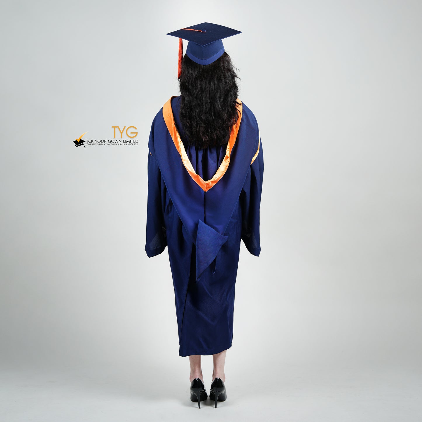A full back view shot of a person in a navy blue graduation gown and an orange hood and with a mortarboard cap with an orange tassel against a gray background.