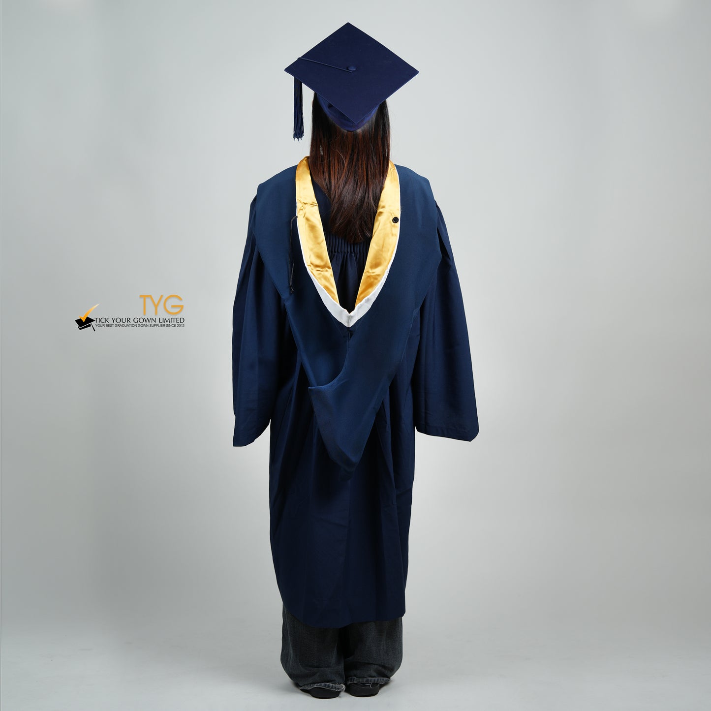 A full body back view of a person in a navy blue graduation gown and a white hood with gold lining and with a mortarboard cap with a white tassel against a gray background.