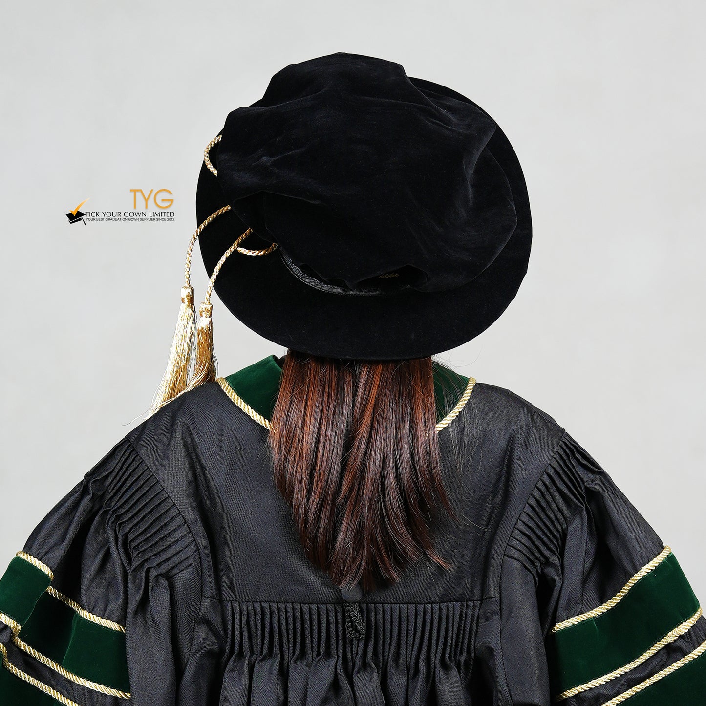 Rear view of a person wearing a black doctoral bonnet and gown with gold accents on a light gray background.