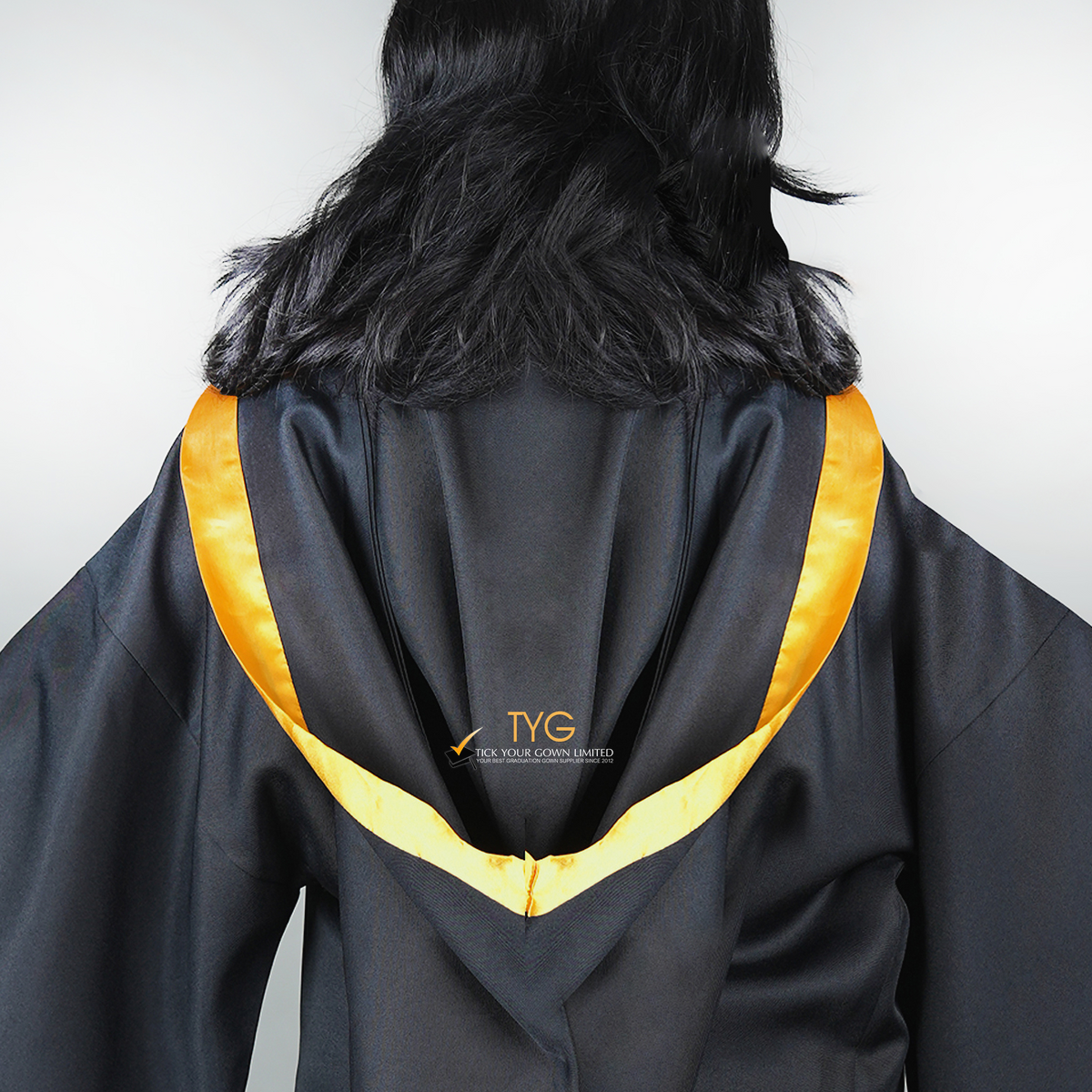 Detailed back view of a graduate in black graduation gown wearing a gold bachelor hood against a plain background