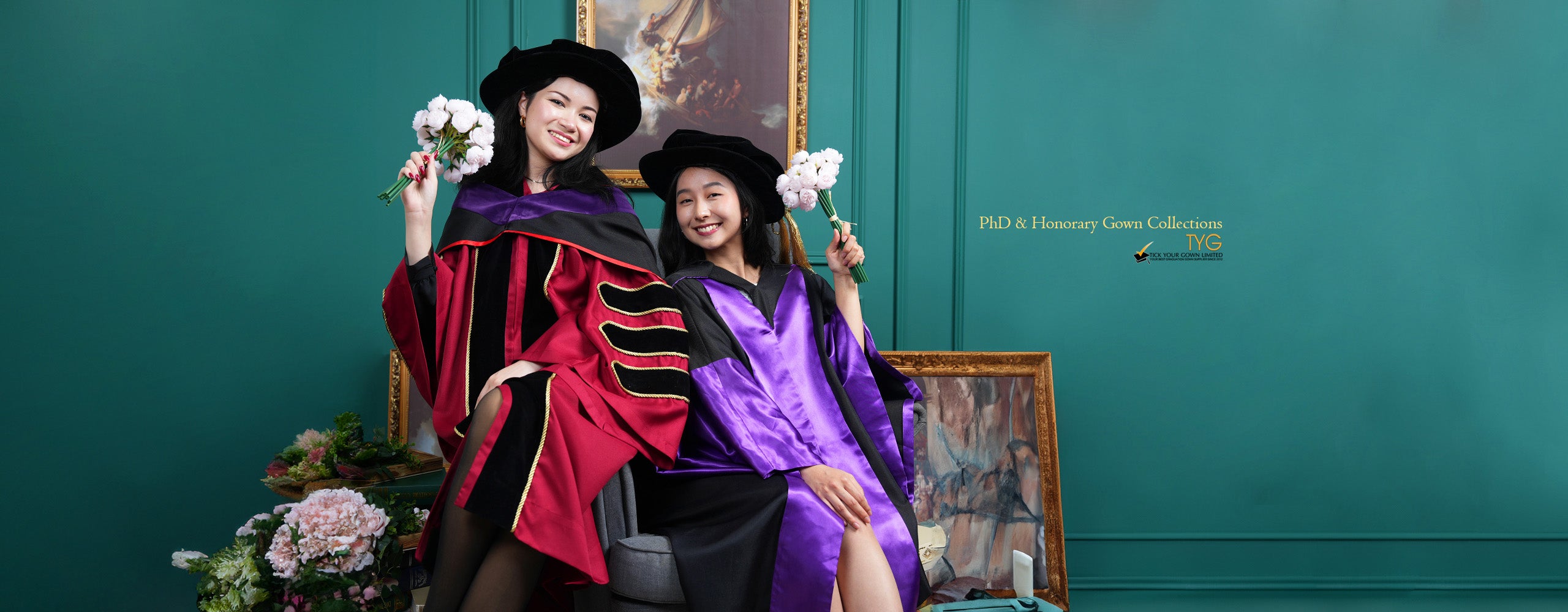 Two individuals in PhD academic regalia holding flowers against a teal wall with framed artwork.