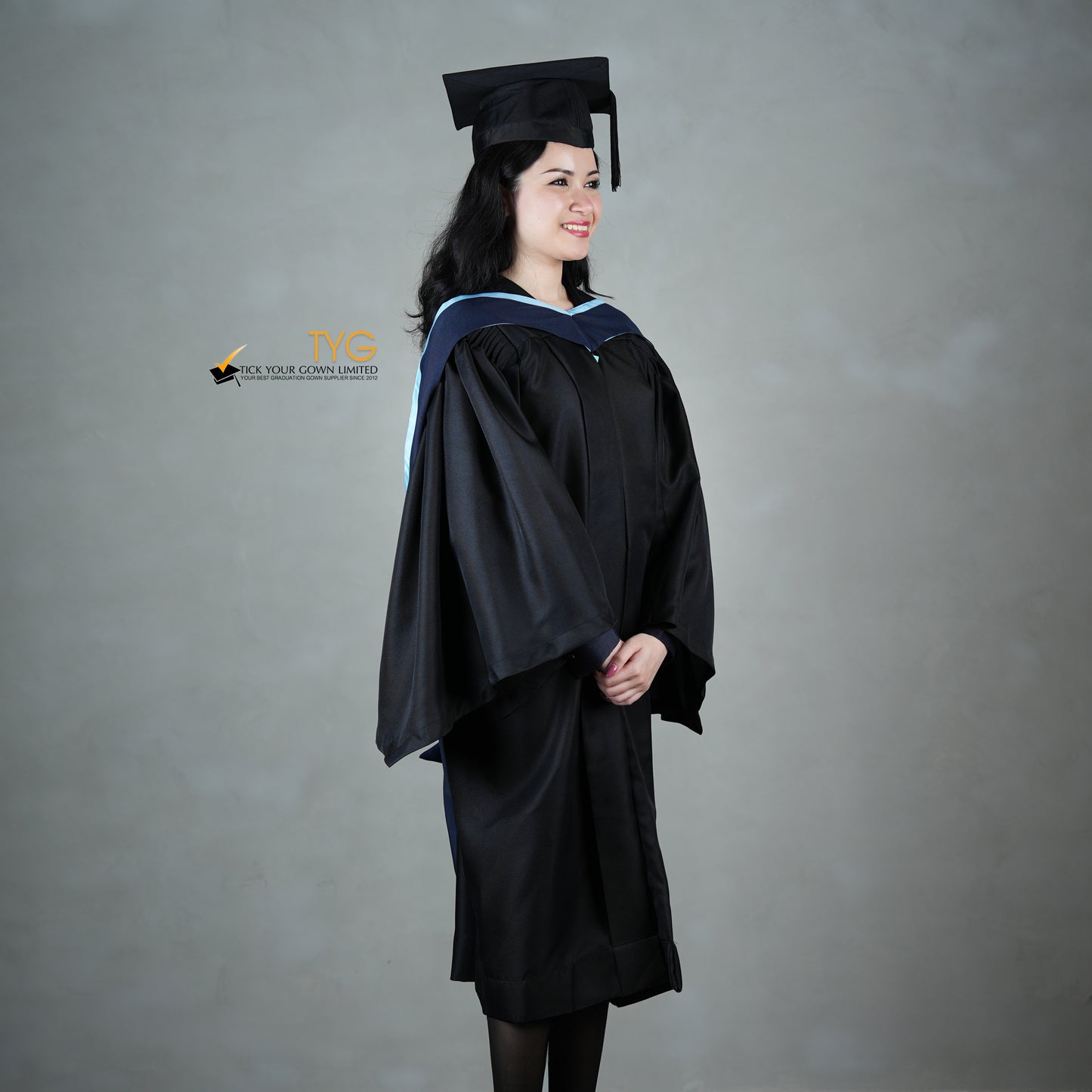 A profile view of a graduate in a black cap and gown with a navy blue hood with light blue lining against a gray background