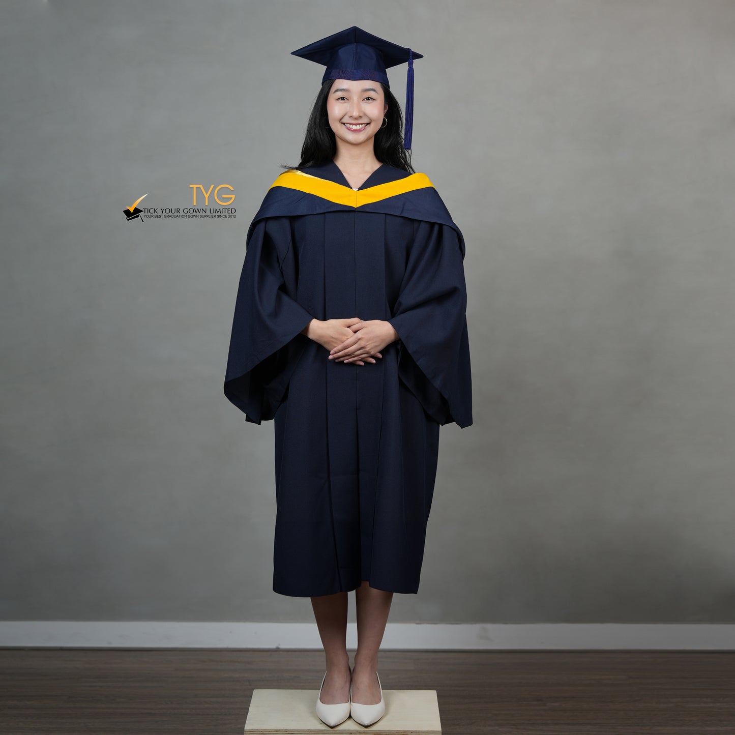 A full front view of a graduate in a navy cap and gown with a yellow hood against a gray background