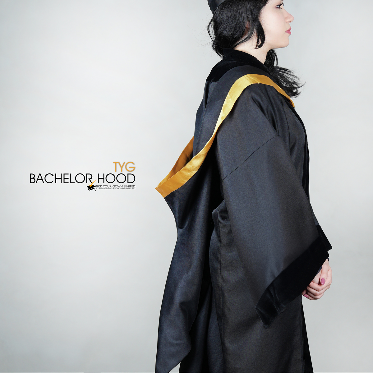 A side view of a graduate in a black graduation gown with a gold bachelor hood against a plain background