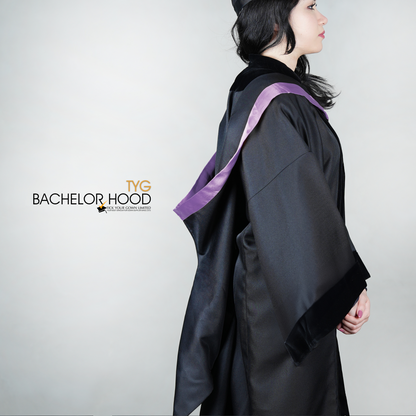 A side view of a graduate in a black graduation gown with a light purple bachelor hood against a plain background
