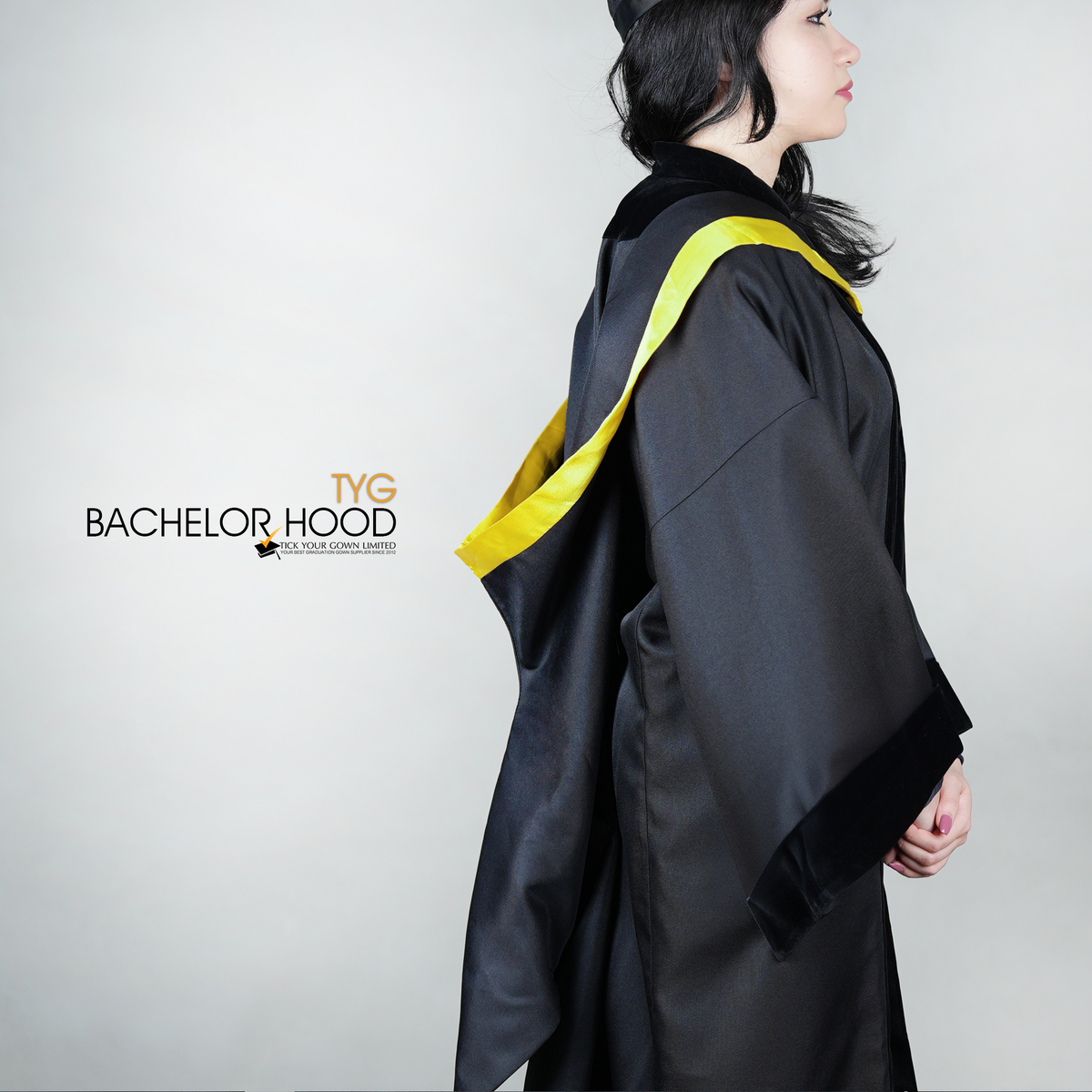 A side view of a graduate in a black graduation gown with a yellow bachelor hood against a plain background