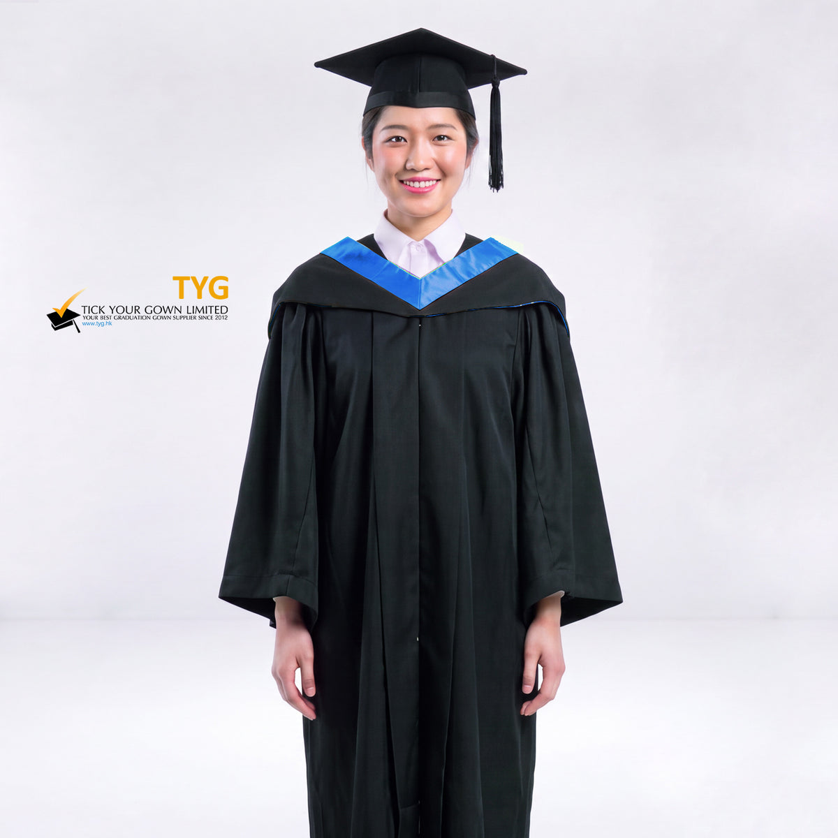 A woman wearing a black graduation gown and a black mortarboard cap stands against a light gray studio background. 

The gown has a blue bachelor hood, and a white collared shirt is visible underneath.
