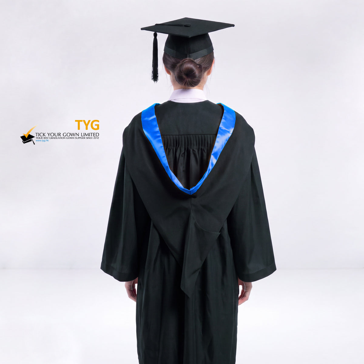 A back view of a woman wearing a black graduation gown and a black mortarboard cap stands against a light gray studio background. 

The gown has a blue bachelor hood, and a white collared shirt is visible underneath.