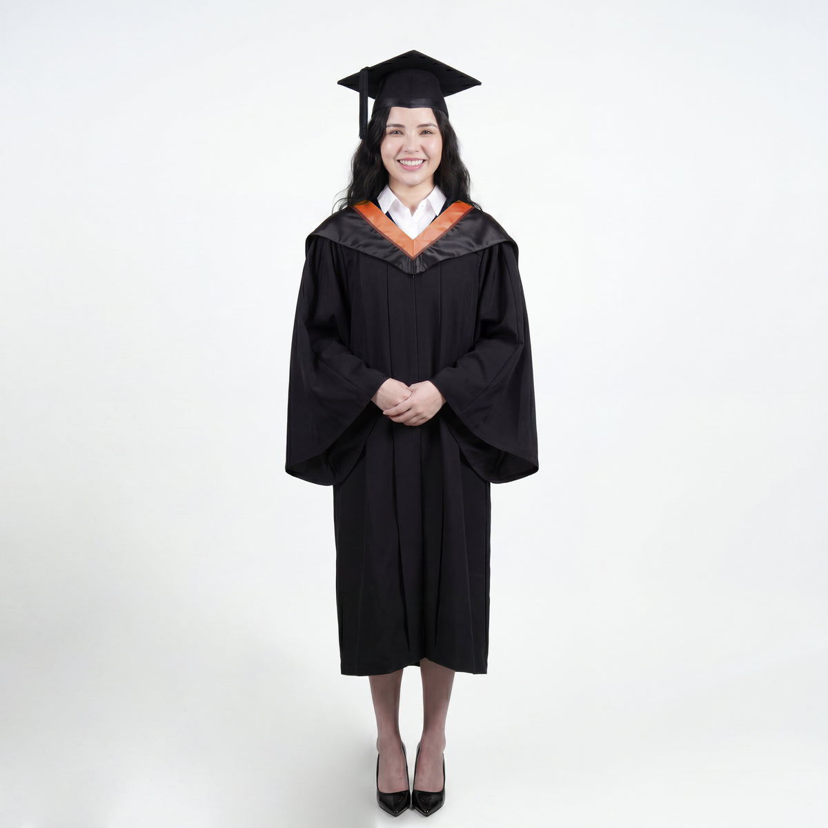 A woman wearing a black graduation gown and matching mortarboard cap stands against a light gray studio background.

The gown has a wide collar with copper and brown trim accent hood, and the model is posed with hands clasped in front.