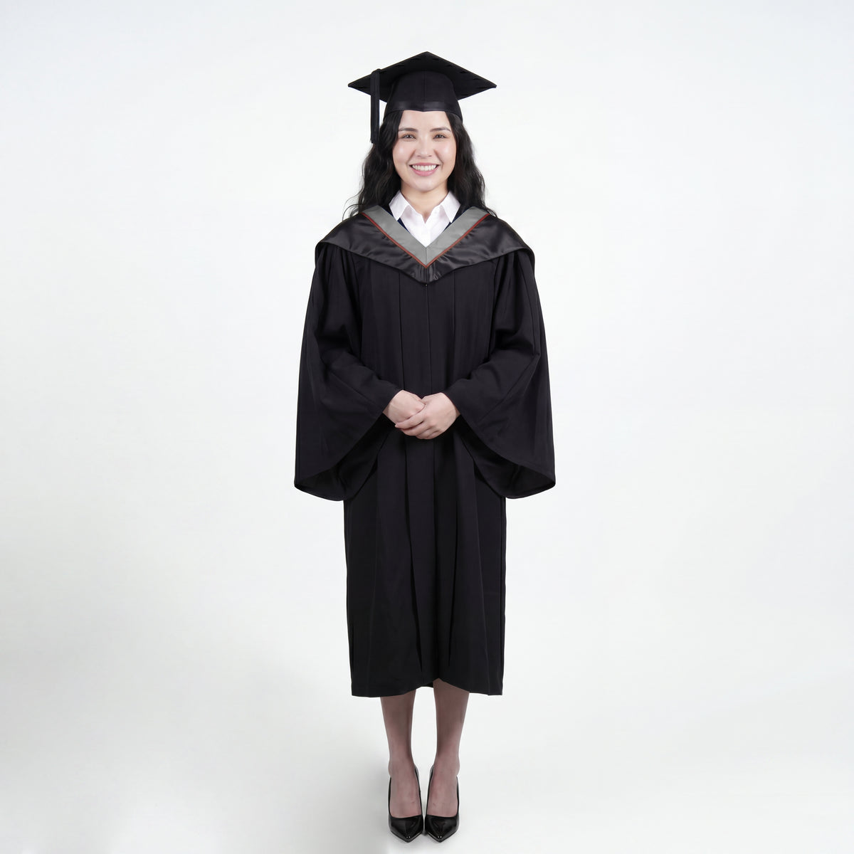 A woman wearing a black graduation gown and matching mortarboard cap stands against a light gray studio background.

The gown has a wide collar with grey and brown trim accent hood, and the model is posed with hands clasped in front.
