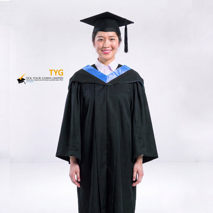 A woman wearing a black graduation gown and a black mortarboard cap stands against a light gray studio background. 

The gown has a light blue bachelor hood, and a white collared shirt is visible underneath.