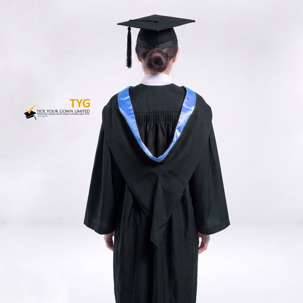 A back view of a woman wearing a black graduation gown and a black mortarboard cap stands against a light gray studio background. 

The gown has a light blue bachelor hood, and a white collared shirt is visible underneath.