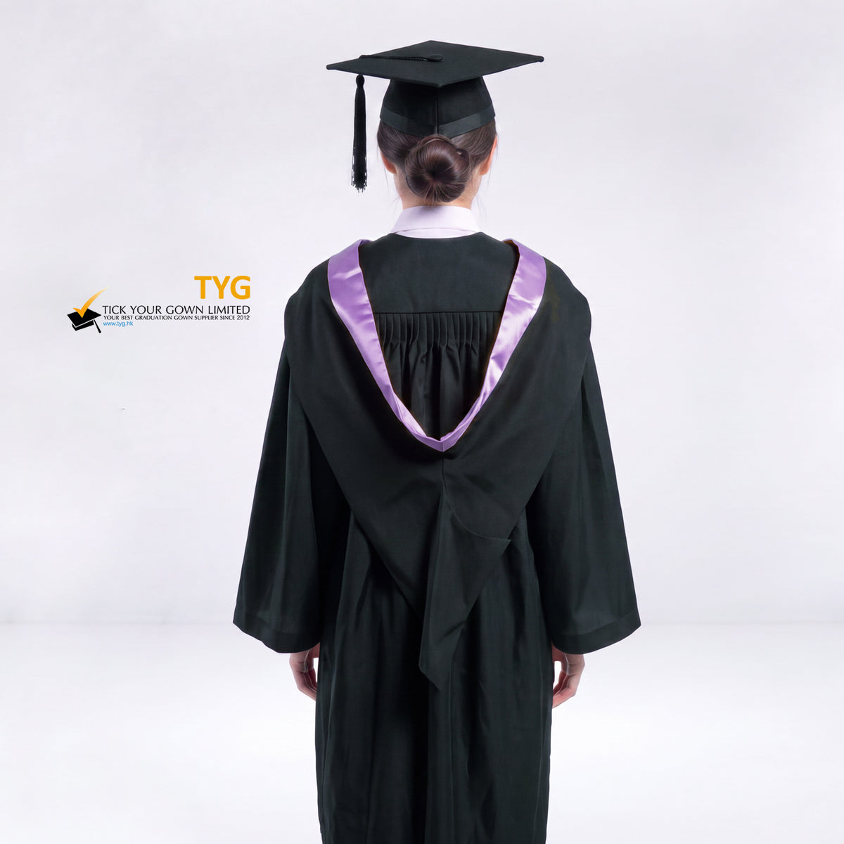 A back view of a woman wearing a black graduation gown and a black mortarboard cap stands against a light gray studio background. 

The gown has a lilac bachelor hood, and a white collared shirt is visible underneath.