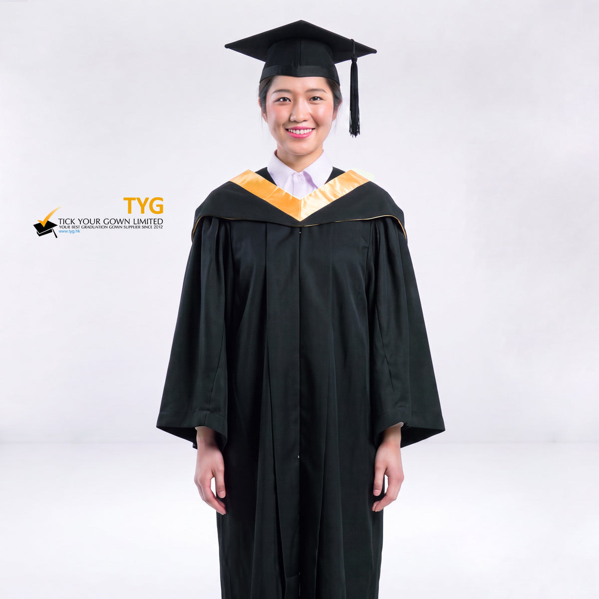 A woman wearing a black graduation gown and a black mortarboard cap stands against a light gray studio background. 

The gown has an orange bachelor hood, and a white collared shirt is visible underneath.