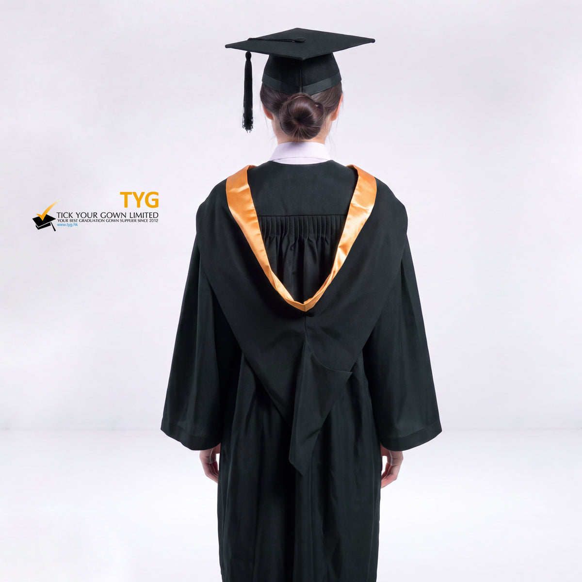 A back view of a woman wearing a black graduation gown and a black mortarboard cap stands against a light gray studio background. 

The gown has an orange bachelor hood, and a white collared shirt is visible underneath.