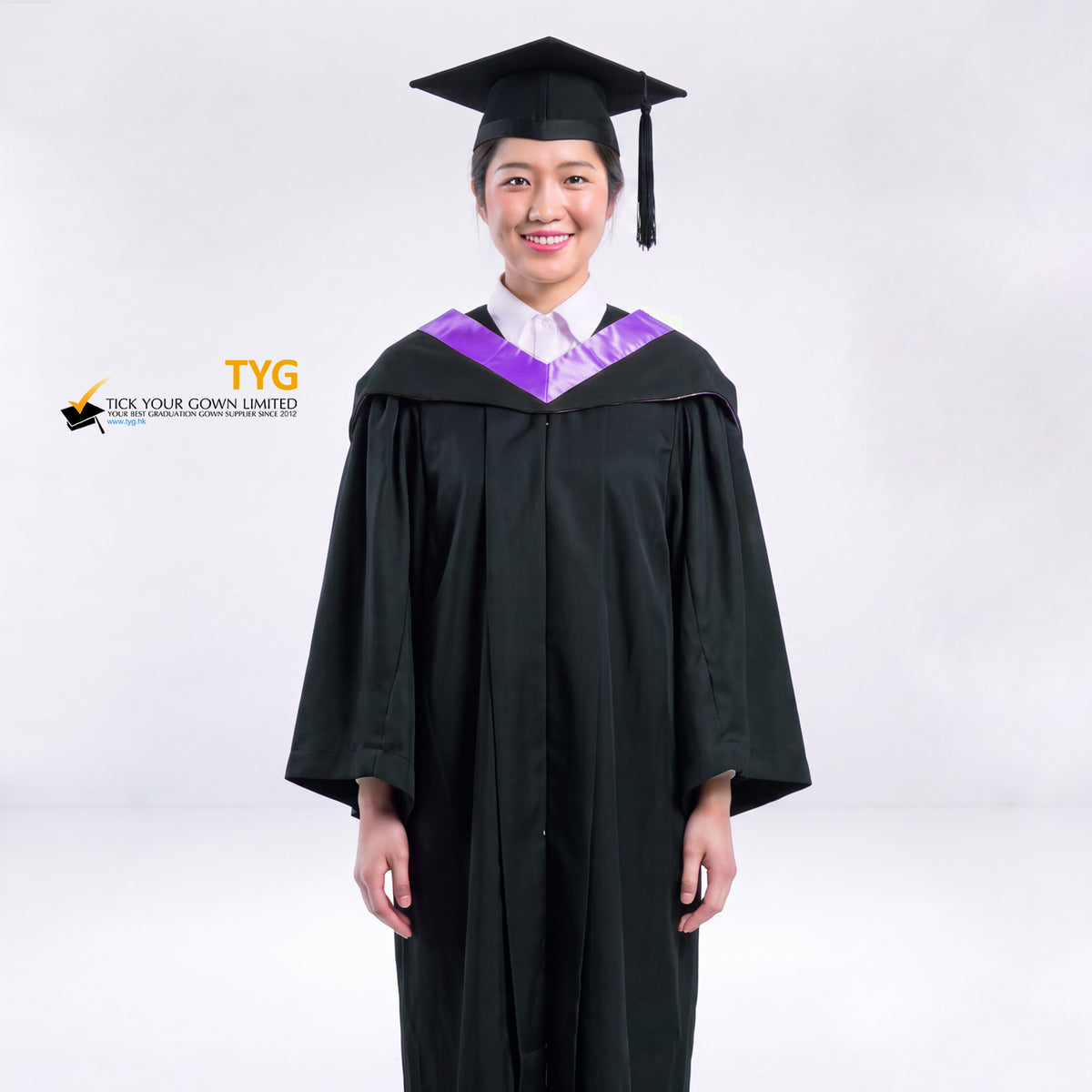 A woman wearing a black graduation gown and a black mortarboard cap stands against a light gray studio background. 

The gown has a purple bachelor hood, and a white collared shirt is visible underneath.