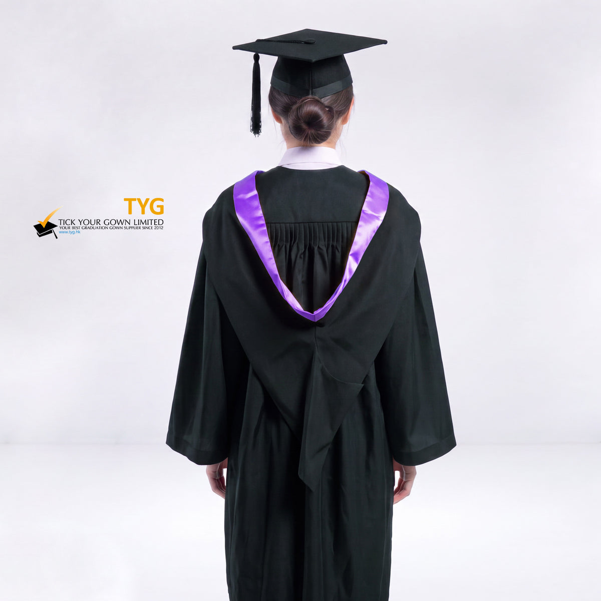 A back view of a woman wearing a black graduation gown and a black mortarboard cap stands against a light gray studio background. 

The gown has a purple bachelor hood, and a white collared shirt is visible underneath.