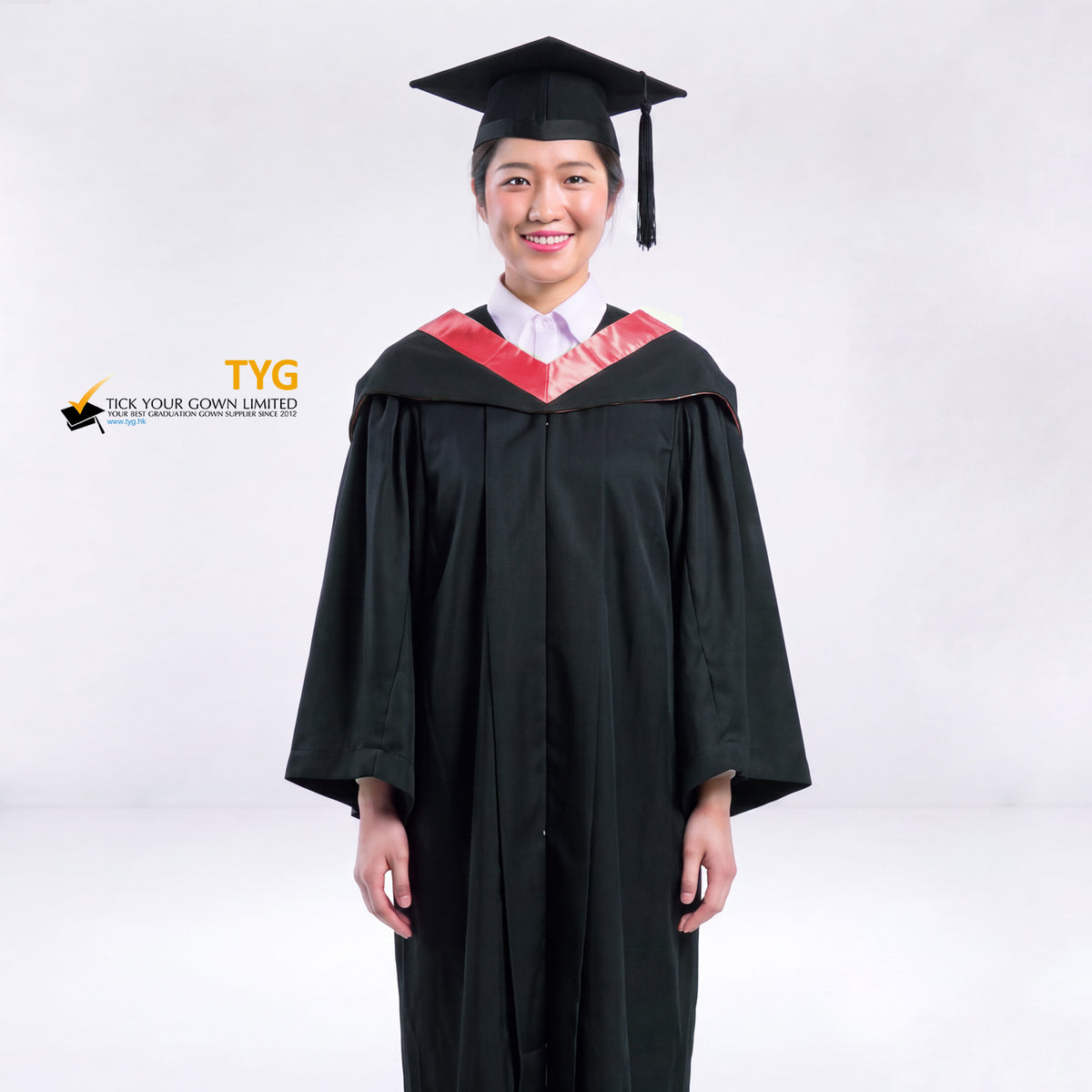 A woman wearing a black graduation gown and a black mortarboard cap stands against a light gray studio background. 

The gown has a red bachelor hood, and a white collared shirt is visible underneath.
