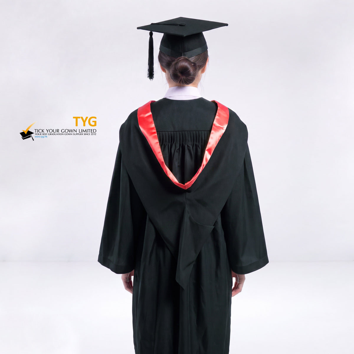 A back view of a woman wearing a black graduation gown and a black mortarboard cap stands against a light gray studio background. 

The gown has a red bachelor hood, and a white collared shirt is visible underneath.