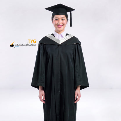 A woman wearing a black graduation gown and a black mortarboard cap stands against a light gray studio background. 

The gown has a silver bachelor hood, and a white collared shirt is visible underneath.