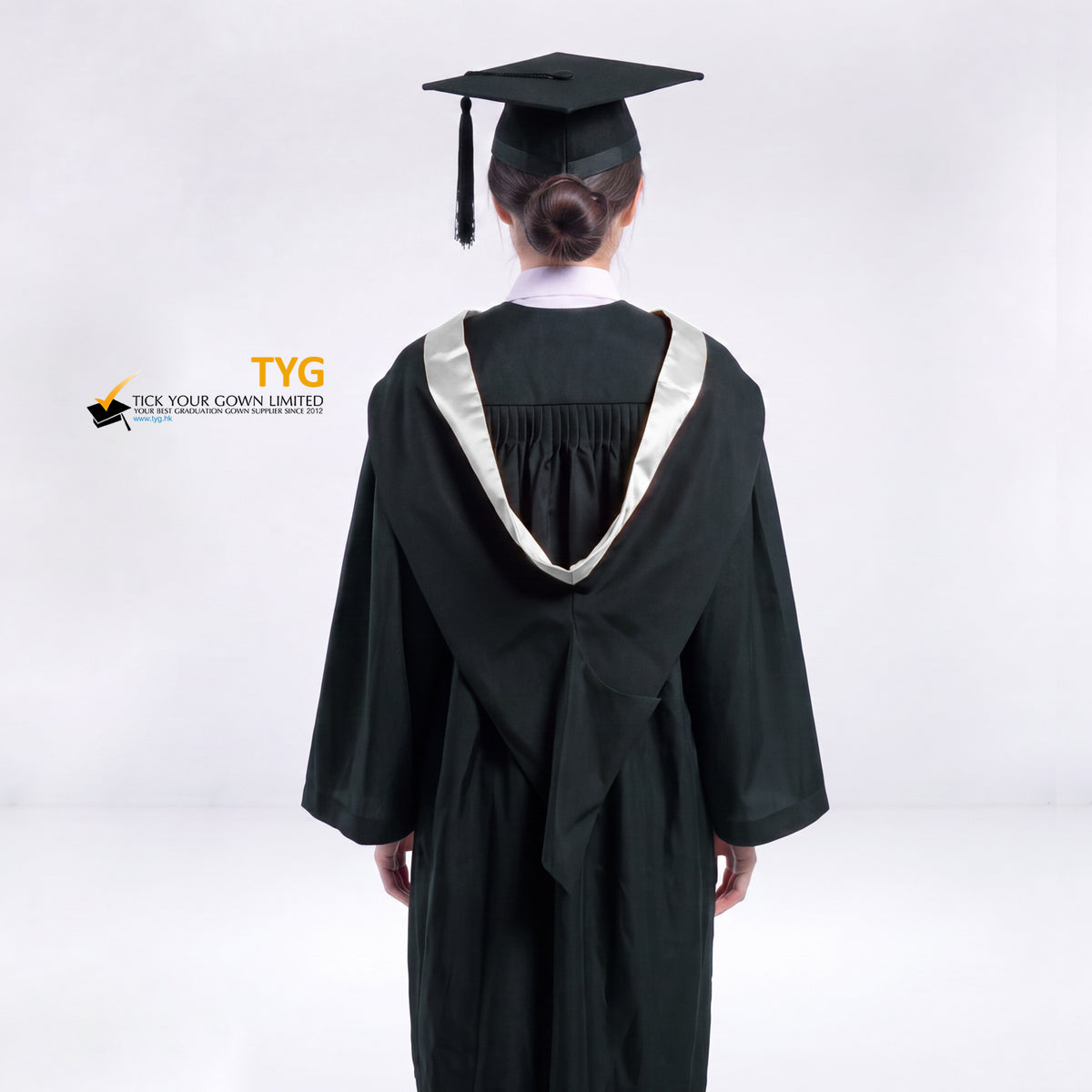 A back view of a woman wearing a black graduation gown and a black mortarboard cap stands against a light gray studio background. 

The gown has a silver bachelor hood, and a white collared shirt is visible underneath.