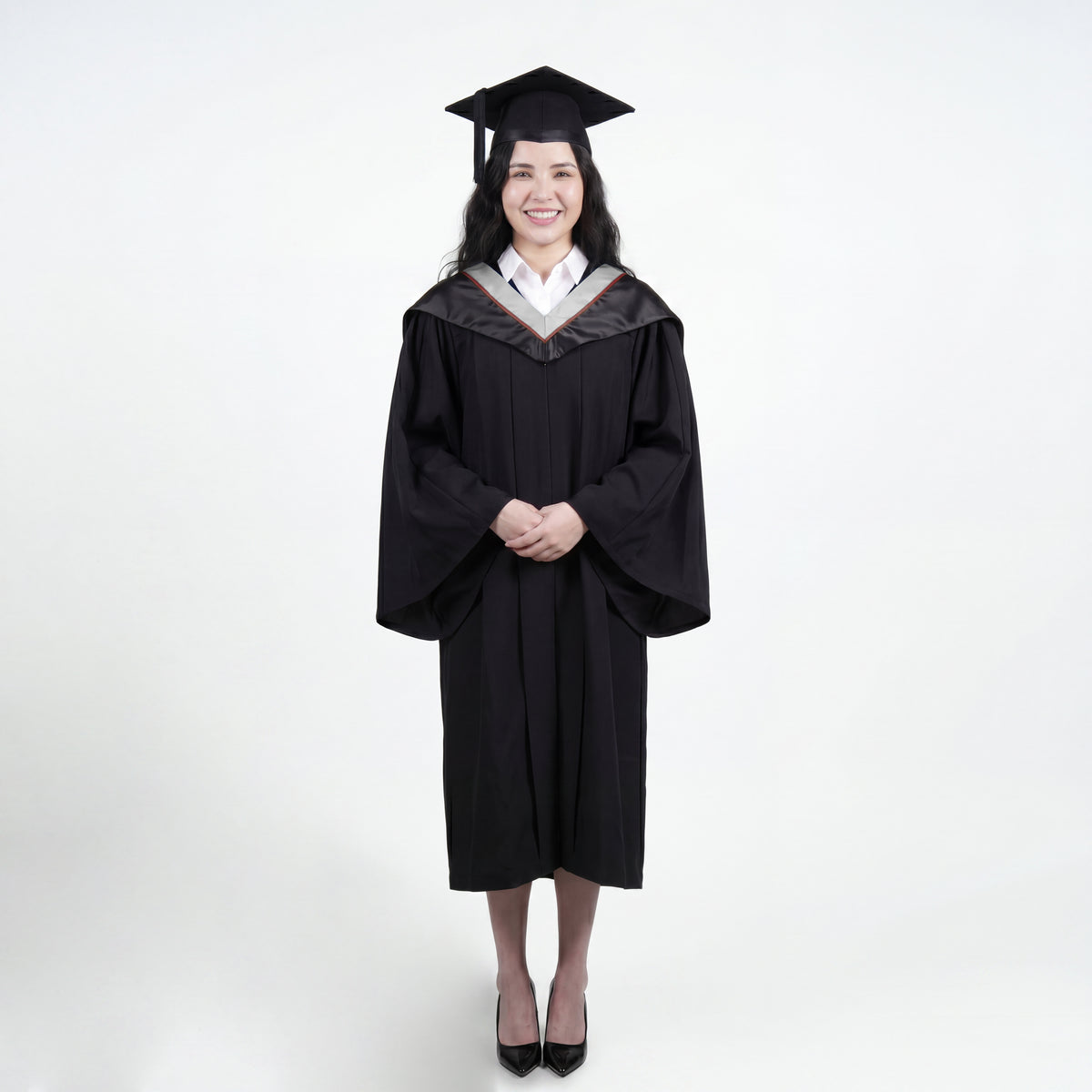 A woman wearing a black graduation gown and matching mortarboard cap stands against a light gray studio background.

The gown has a wide collar with silver and brown trim accent hood, and the model is posed with hands clasped in front.