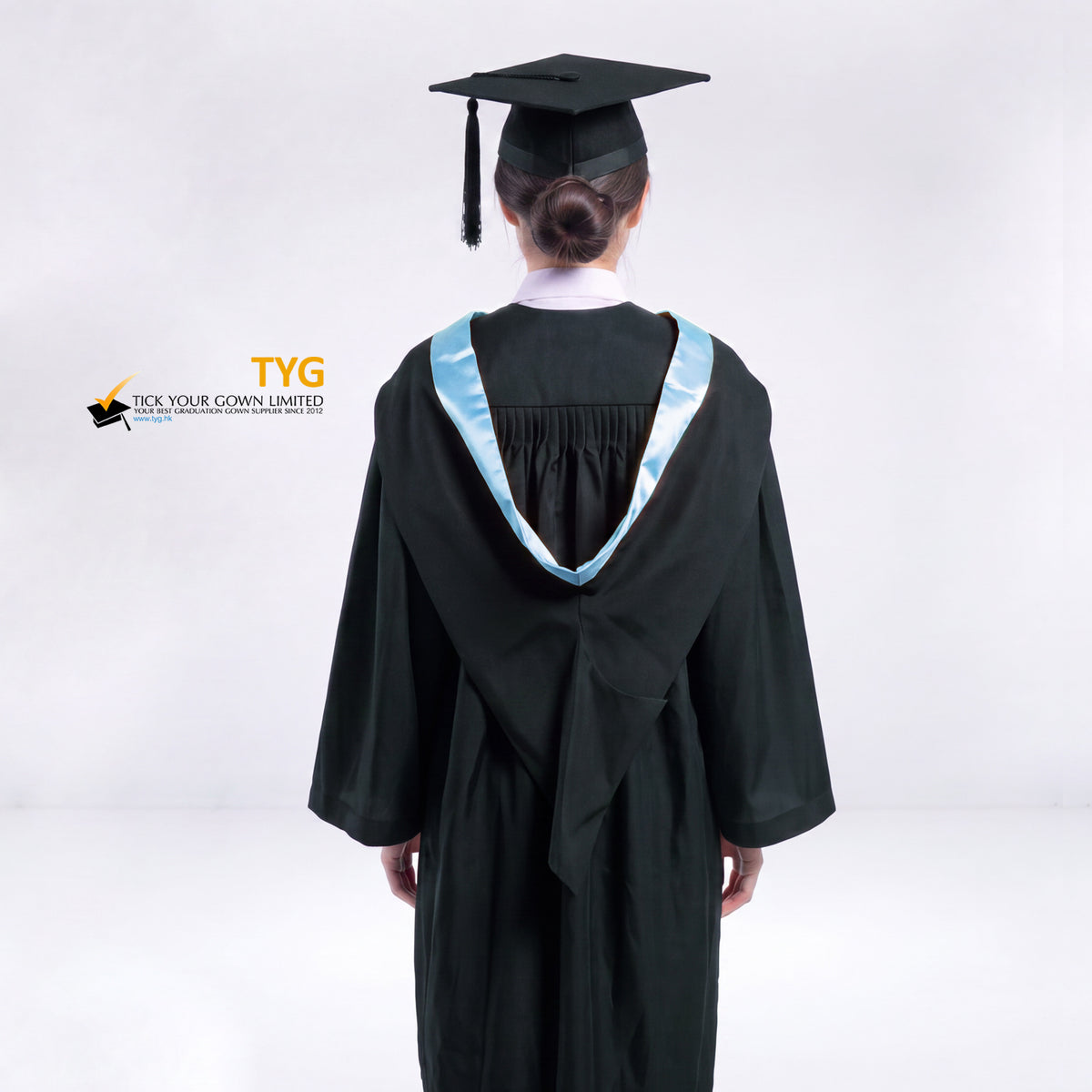 A back view of a woman wearing a black graduation gown and a black mortarboard cap stands against a light gray studio background. 

The gown has a sky blue bachelor hood, and a white collared shirt is visible underneath.