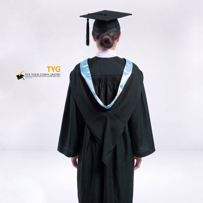 A back view of a woman wearing a black graduation gown and a black mortarboard cap stands against a light gray studio background. 

The gown has a sky blue bachelor hood, and a white collared shirt is visible underneath.