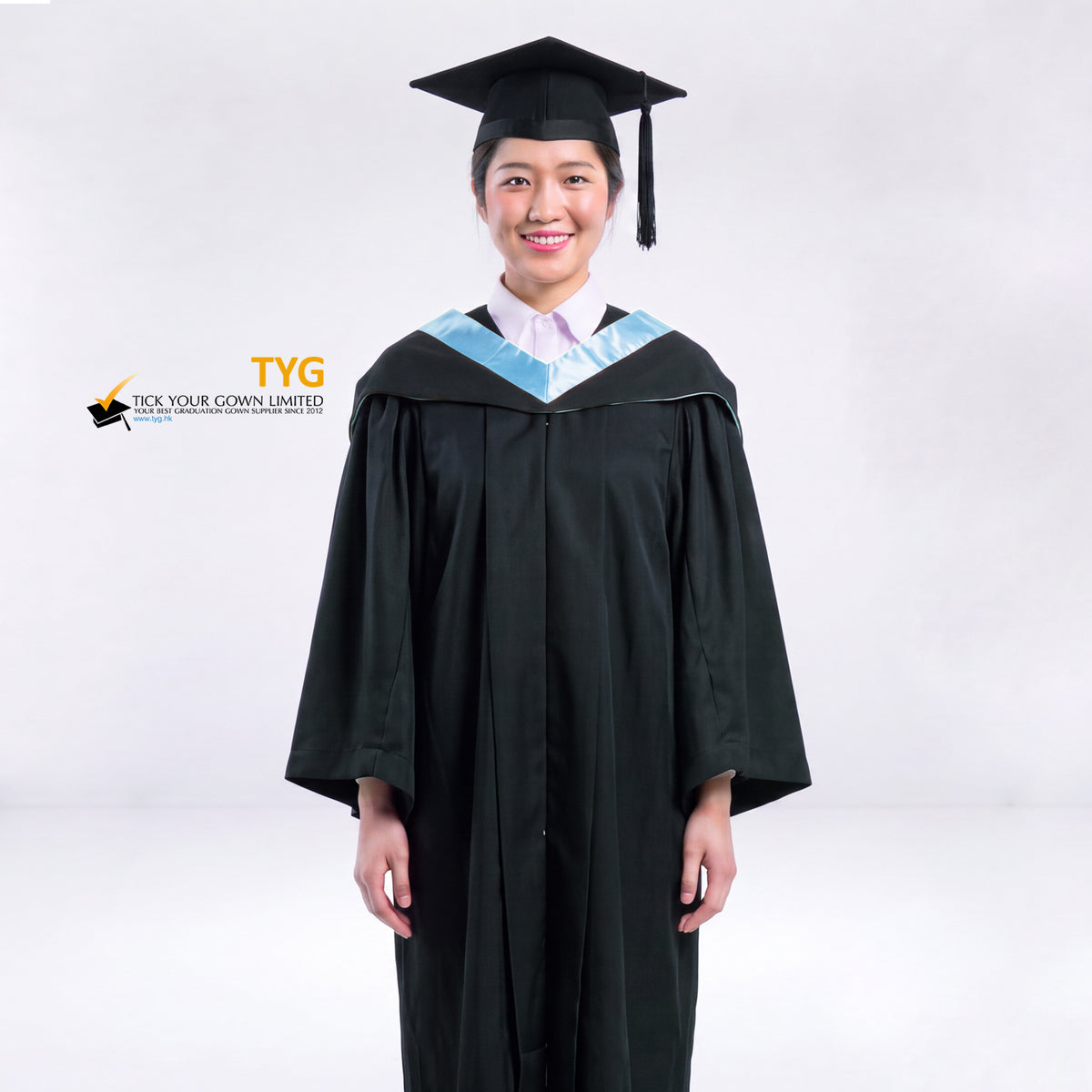 A woman wearing a black graduation gown and a black mortarboard cap stands against a light gray studio background. 

The gown has a sky blue bachelor hood, and a white collared shirt is visible underneath.