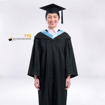 A woman wearing a black graduation gown and a black mortarboard cap stands against a light gray studio background. 

The gown has a sky blue bachelor hood, and a white collared shirt is visible underneath.