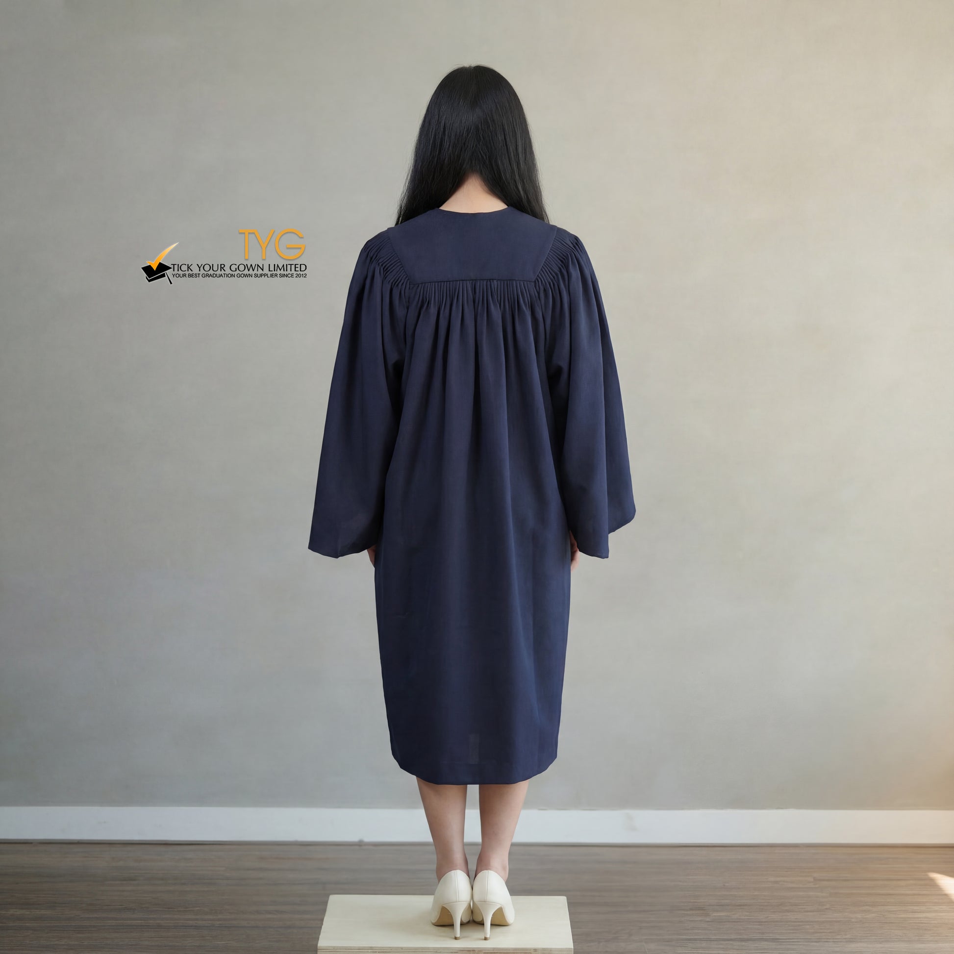 A back view of a person wearing a bachelors graduation gown in navy blue standing on a wooden platform against a plain wall.