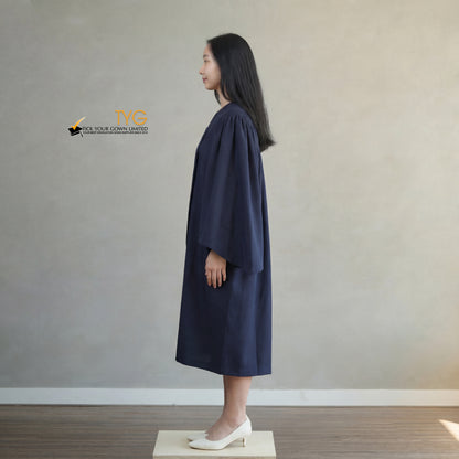 A side view of a person wearing a bachelors graduation gown in navy blue standing on a wooden platform against a plain wall.