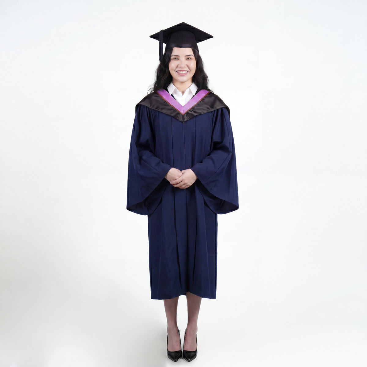 A woman wearing a navy blue graduation gown and matching mortarboard cap stands against a light gray studio background.

The gown has a wide collar with pink and brown trim accent hood, and the model is posed with hands clasped in front.