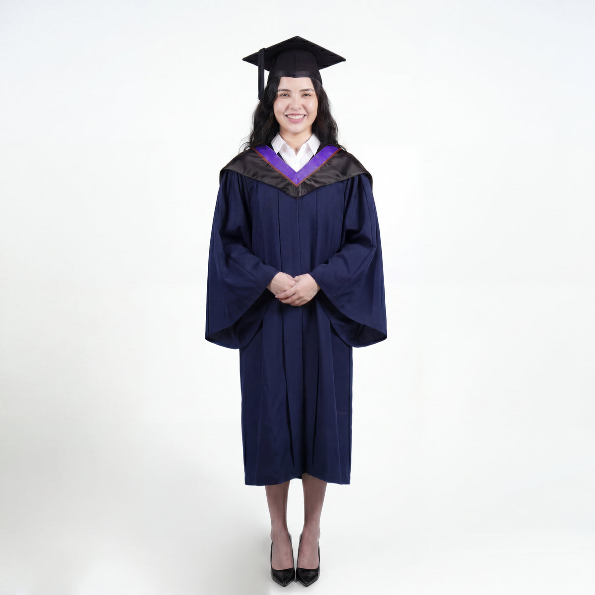 A woman wearing a navy blue graduation gown and matching mortarboard cap stands against a light gray studio background.

The gown has a wide collar with purple and brown trim accent hood, and the model is posed with hands clasped in front.
