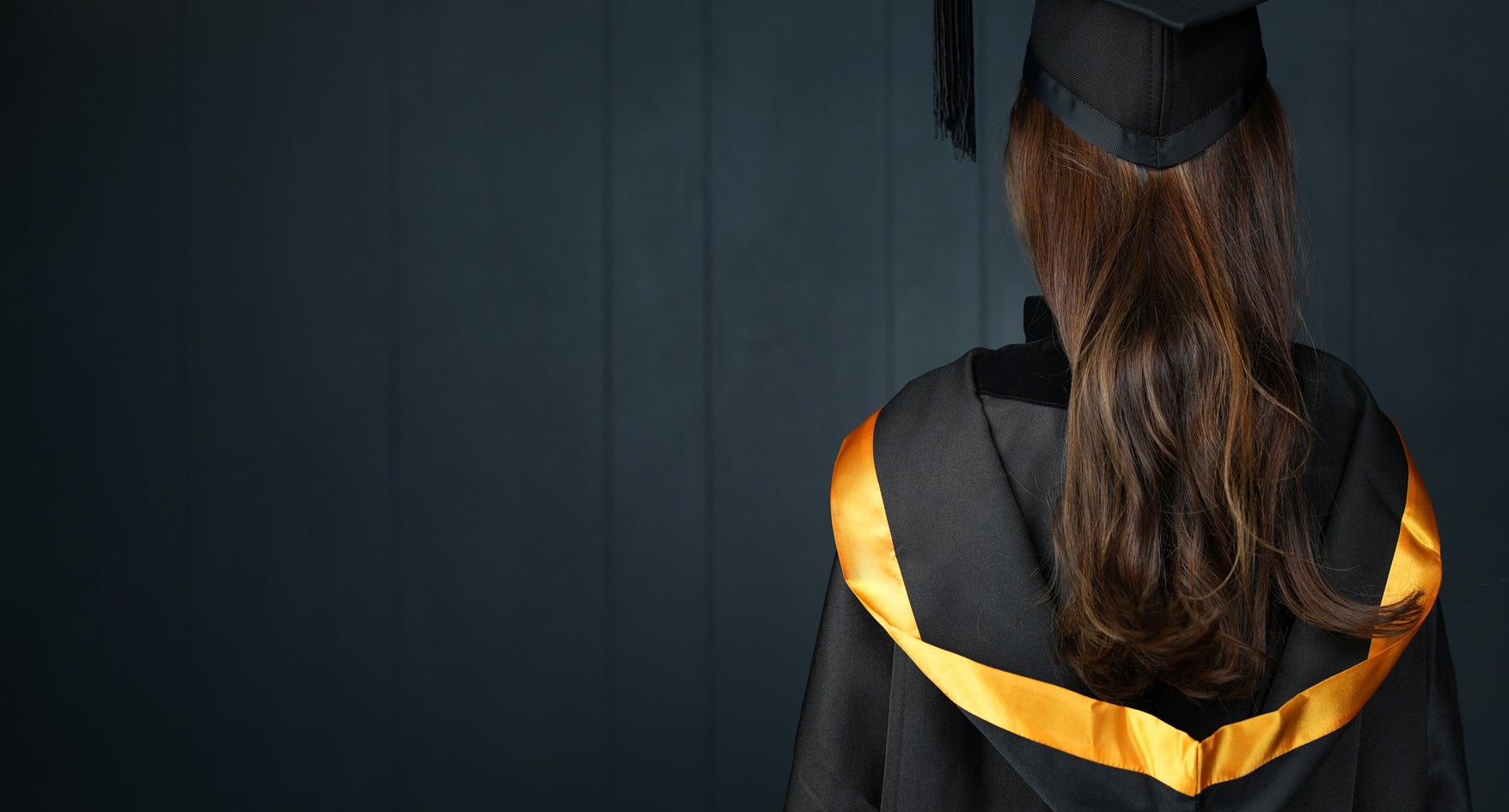 A back view of a person wearing a graduation gown and cap with orange accents on a dark background
