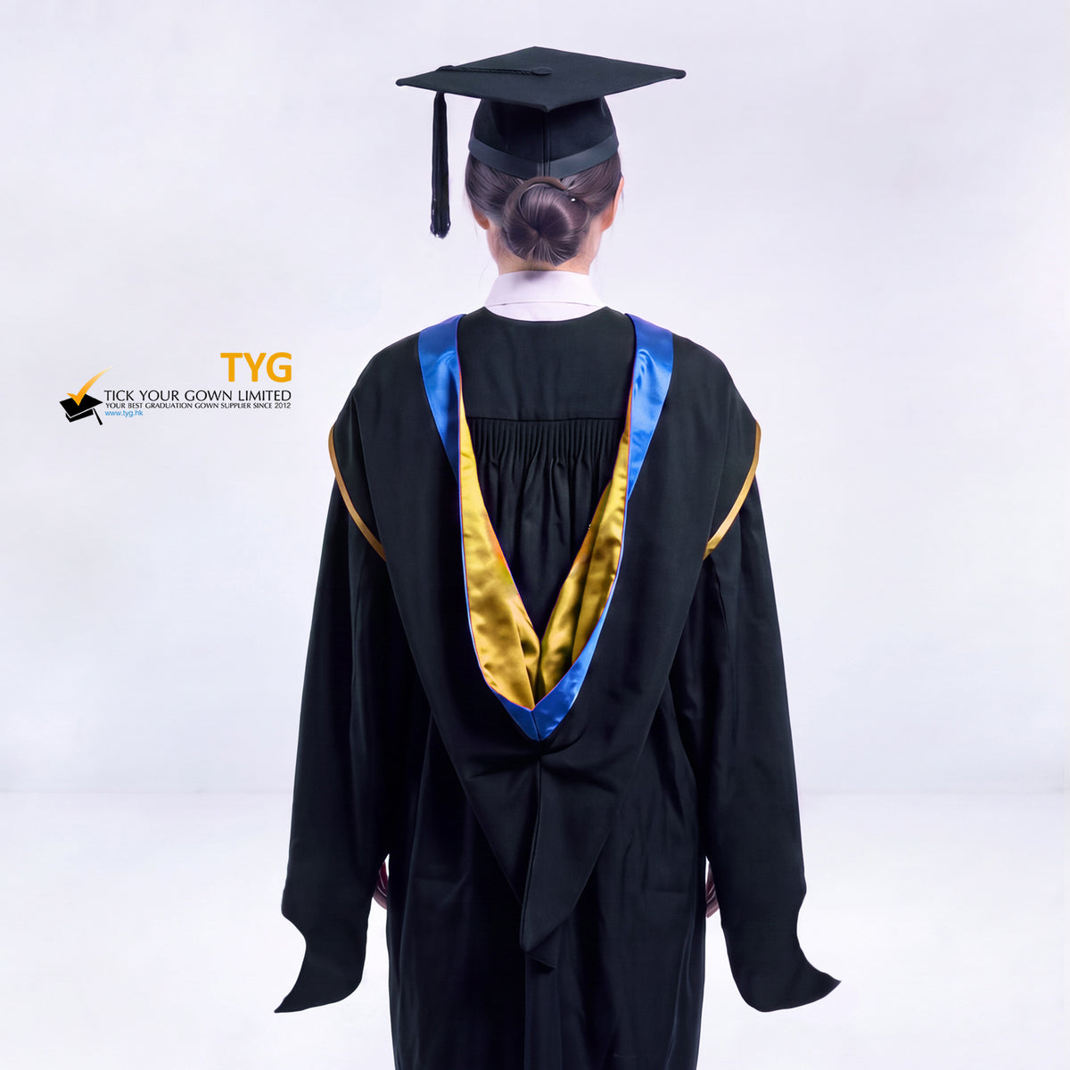 A back view of a woman in a black master's graduation gown classic UK with a blue hood with gold trim accent and with a black mortarboard cap with a black tassel against a gray background.