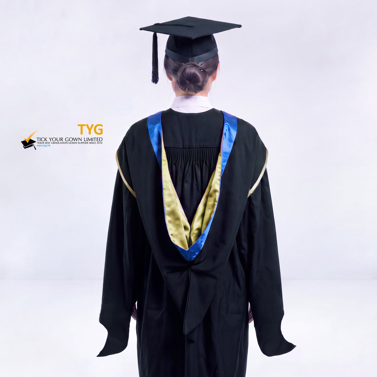 A back view of a woman in a black master's graduation gown classic UK with a blue hood with yellow trim accent and with a black mortarboard cap with a black tassel against a gray background.