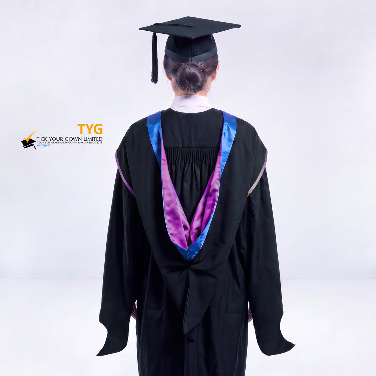 A back view of a woman in a black master's graduation gown classic UK with a blue hood with light purple trim accent and with a black mortarboard cap with a black tassel against a gray background.