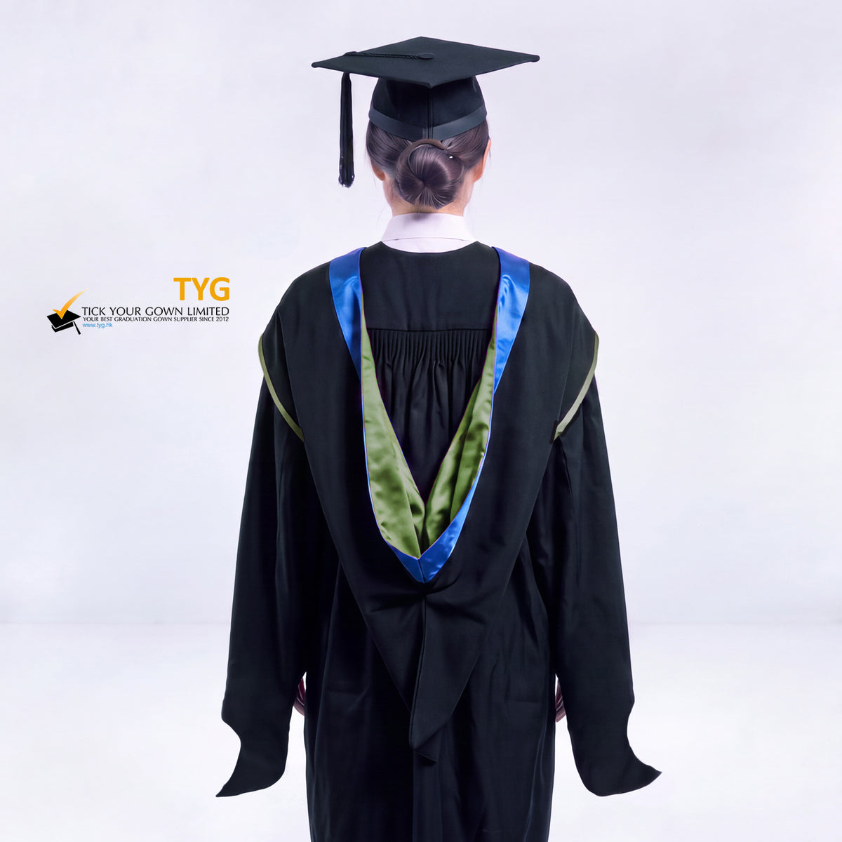 A back view of a woman in a black master's graduation gown classic UK with a blue hood with green trim accent and with a black mortarboard cap with a black tassel against a gray background.