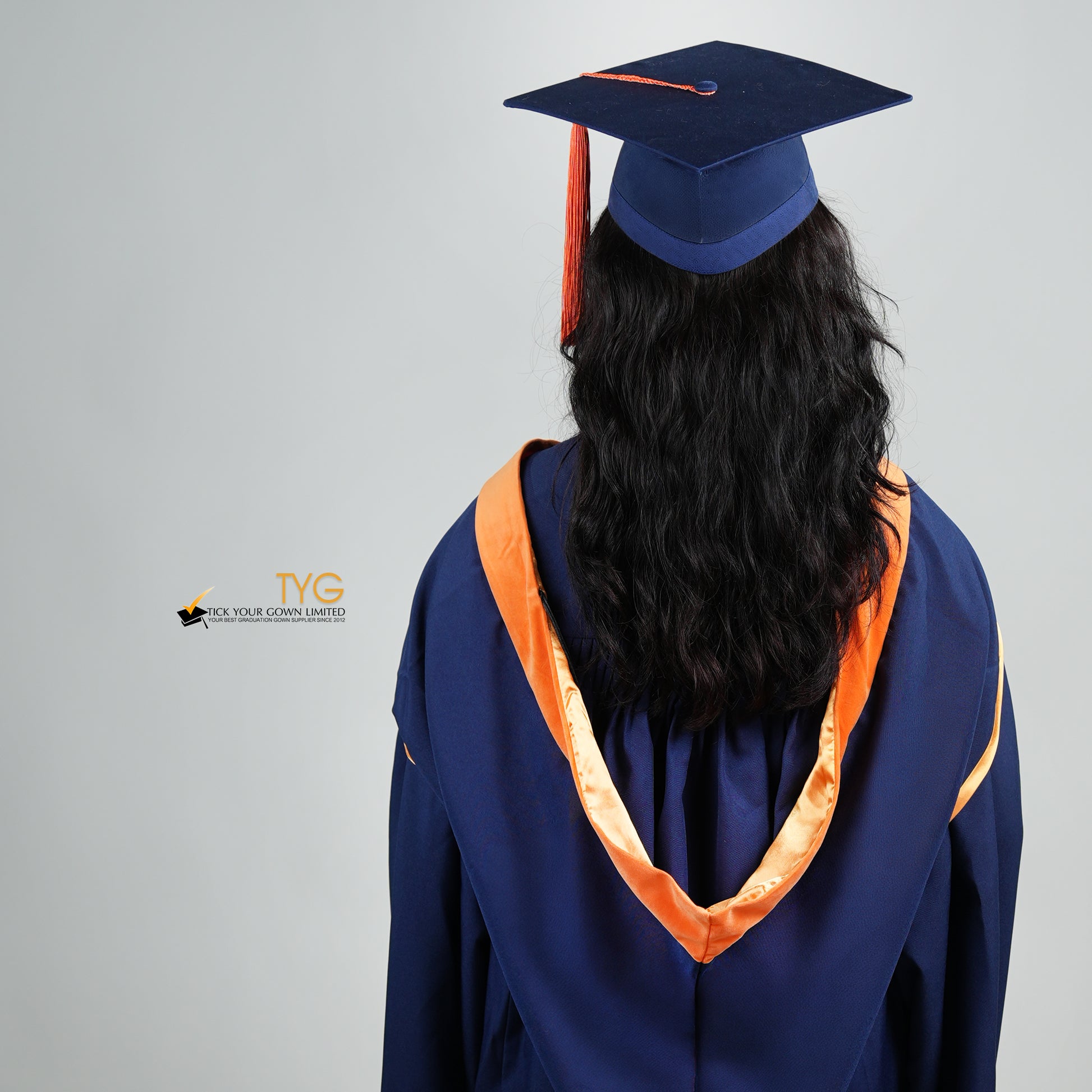 A back view of a person in a navy blue graduation gown and an orange hood and with a mortarboard cap with an orange tassel against a gray background.