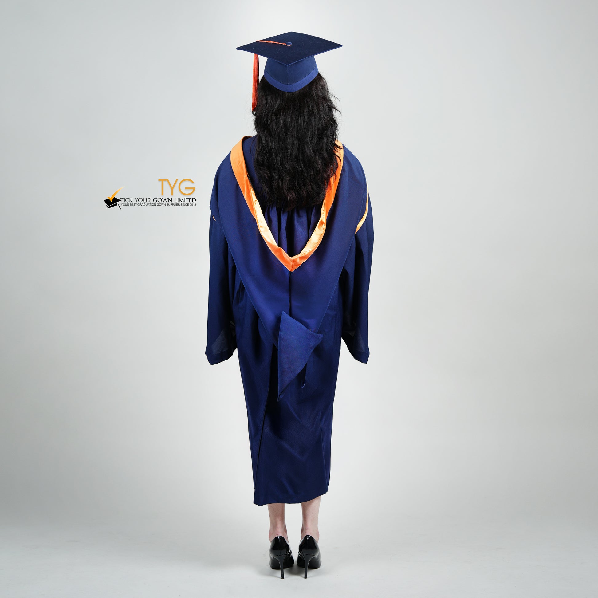 A full back view shot of a person in a navy blue graduation gown and an orange hood and with a mortarboard cap with an orange tassel against a gray background.