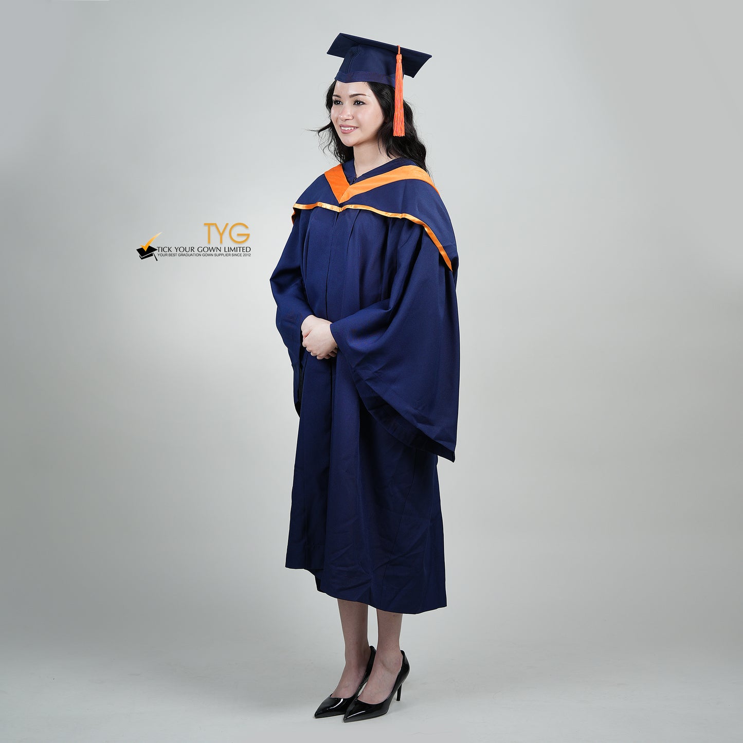 A full body profile shot of a person in a navy blue graduation gown and an orange hood and with a mortarboard cap with an orange tassel against a gray background.