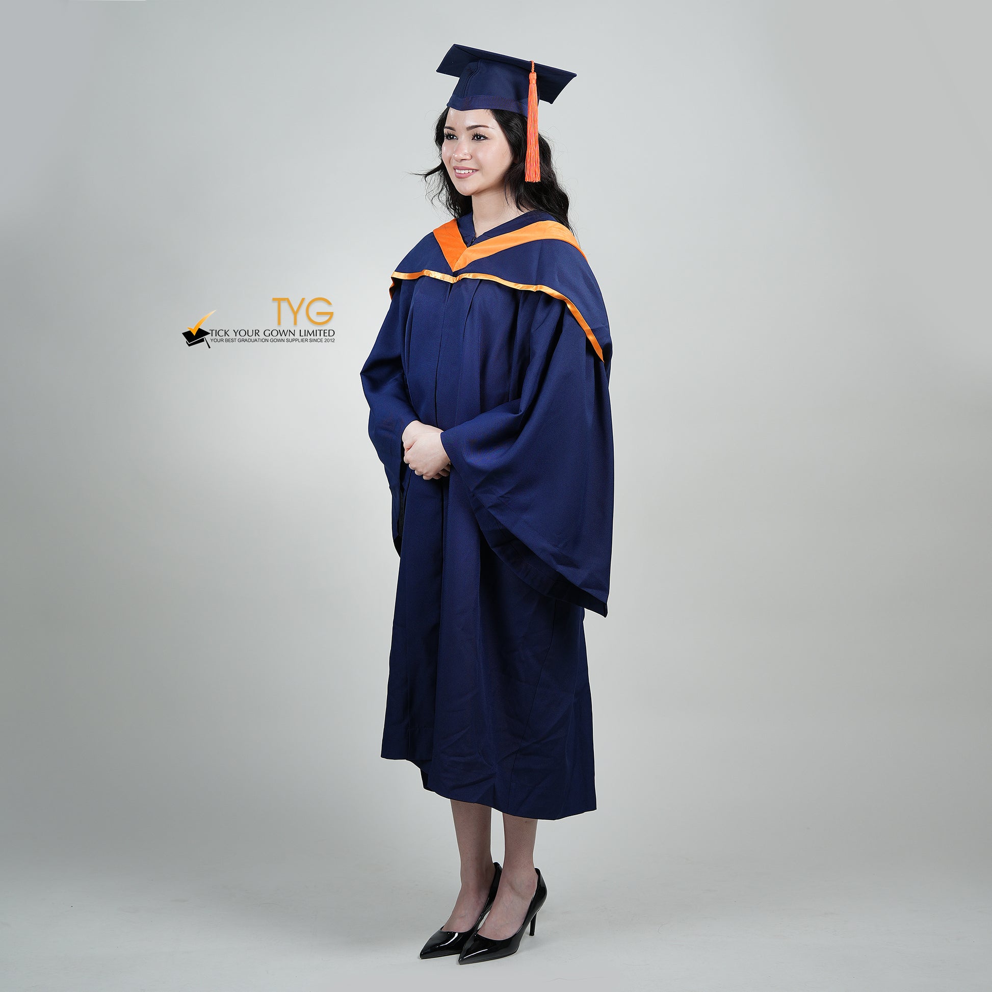 A full body profile shot of a person in a navy blue graduation gown and an orange hood and with a mortarboard cap with an orange tassel against a gray background.
