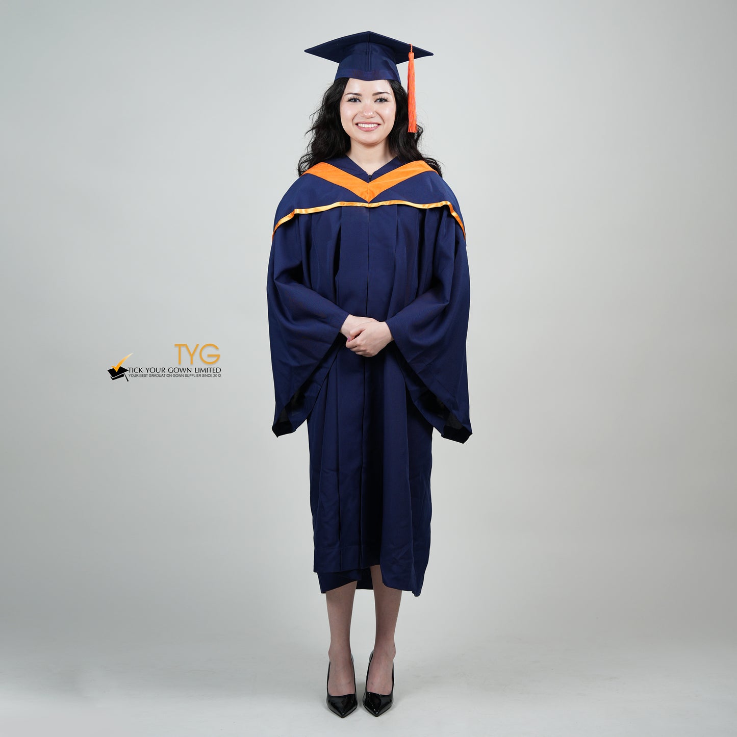 A full body shot of a person in a navy blue graduation gown and an orange hood and with a mortarboard cap with an orange tassel against a gray background.